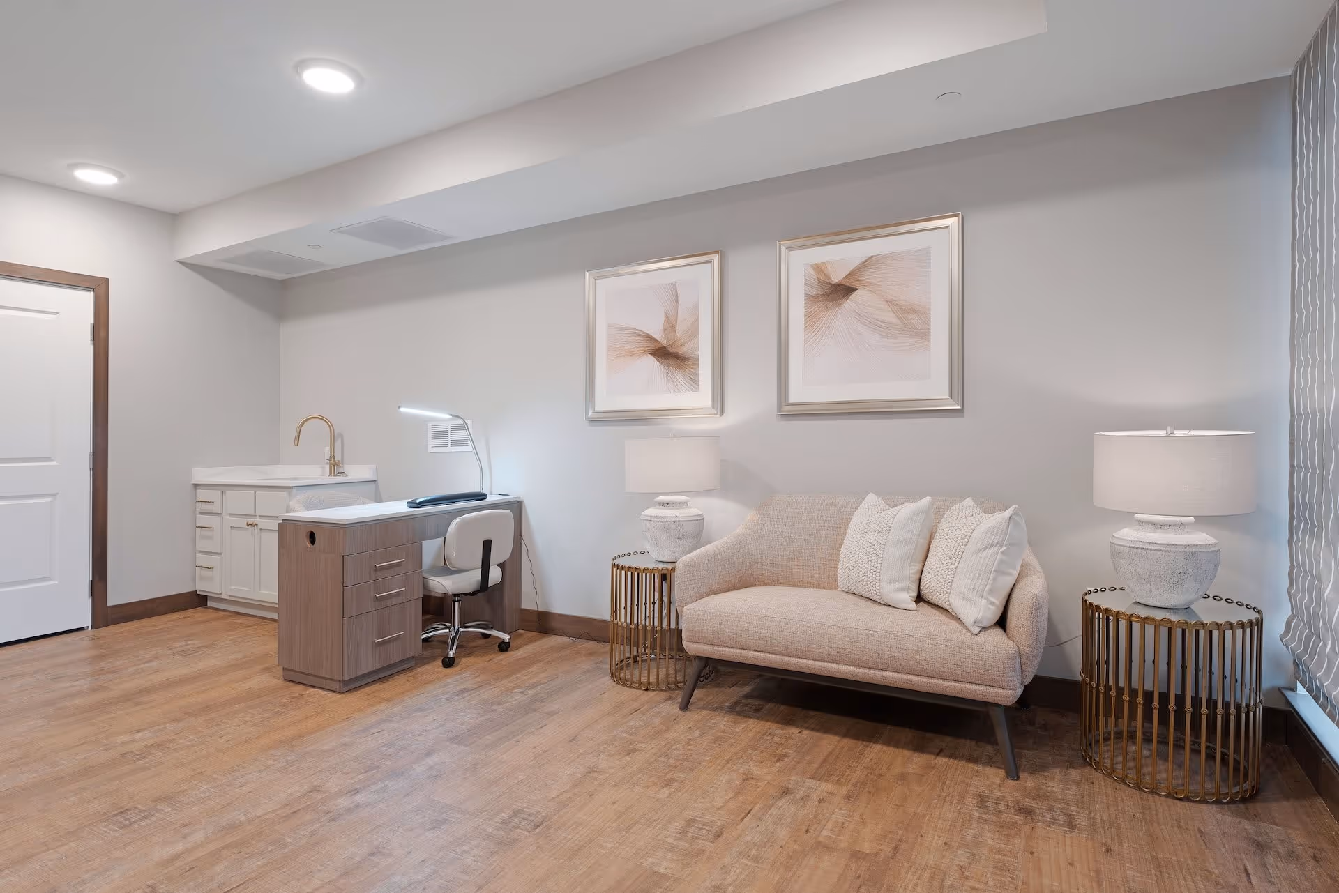 A cozy interior room with wooden flooring featuring a small beige loveseat with three white pillows, two framed abstract artworks on the wall above it, a round side table with a white lamp, and a small desk with a white chair and a desk lamp next to a white cabinet with a gold faucet.