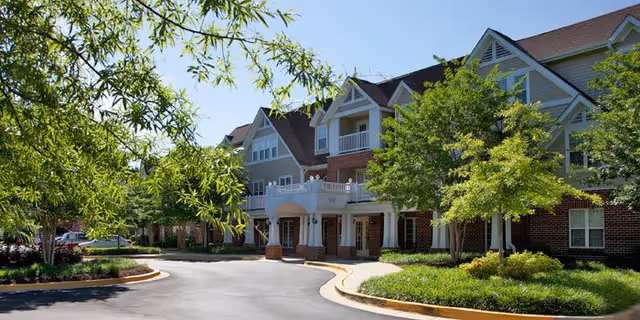 Exterior view of a senior living facility named The Willows, showing a large multi-story building with a combination of brick and siding, surrounded by green trees and landscaping under a clear blue sky.