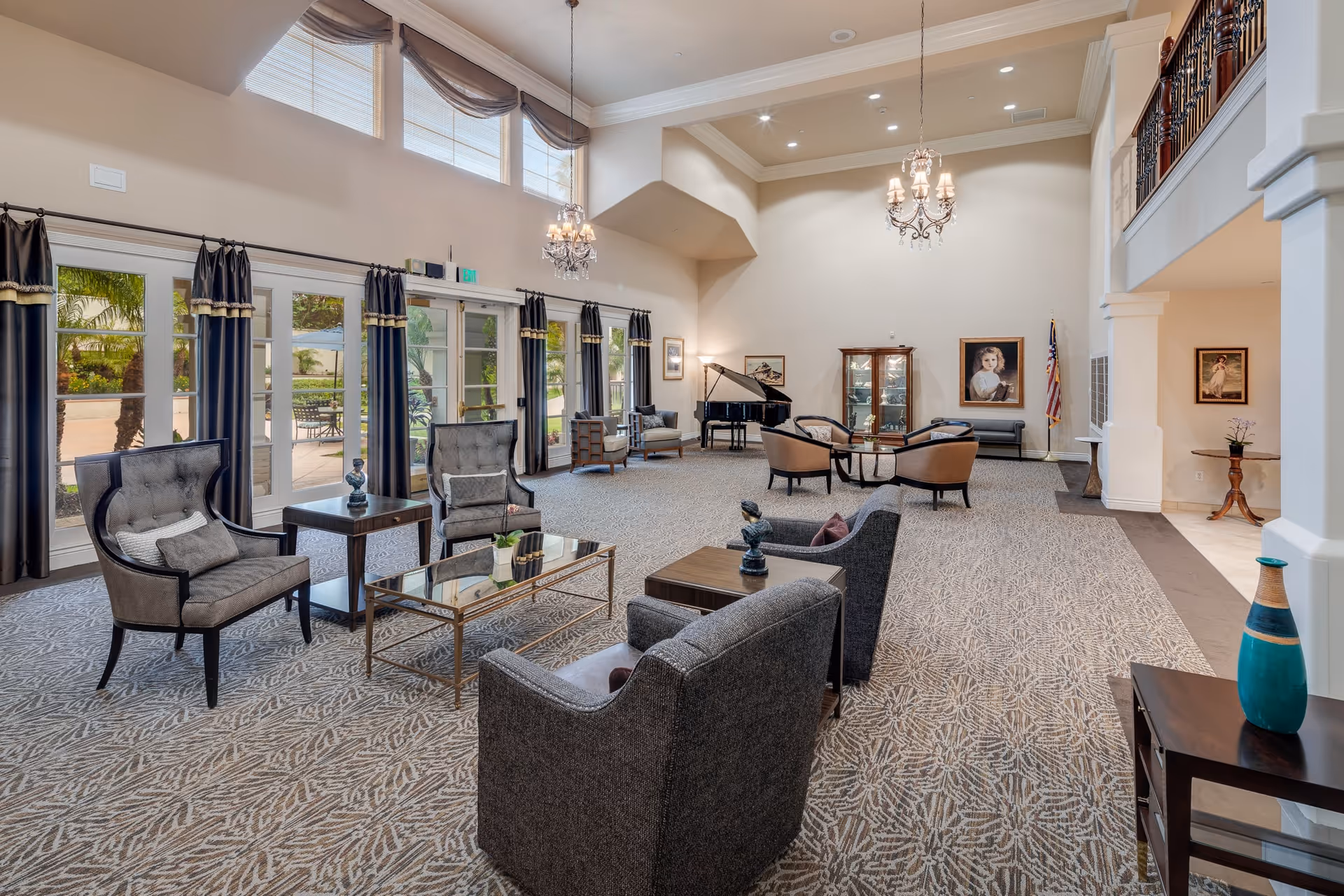 Spacious and elegant living room area in a senior living facility with high ceilings, large windows with dark curtains, multiple seating arrangements including armchairs and sofas, a grand piano, chandeliers, framed portraits on the walls, and decorative vases on tables.