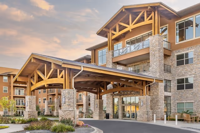 Exterior view of Legacy Village of Stillwater, showing a large covered entrance with wooden beams and stone pillars, attached to a multi-story building with numerous windows and balconies under a partly cloudy sky at sunset.