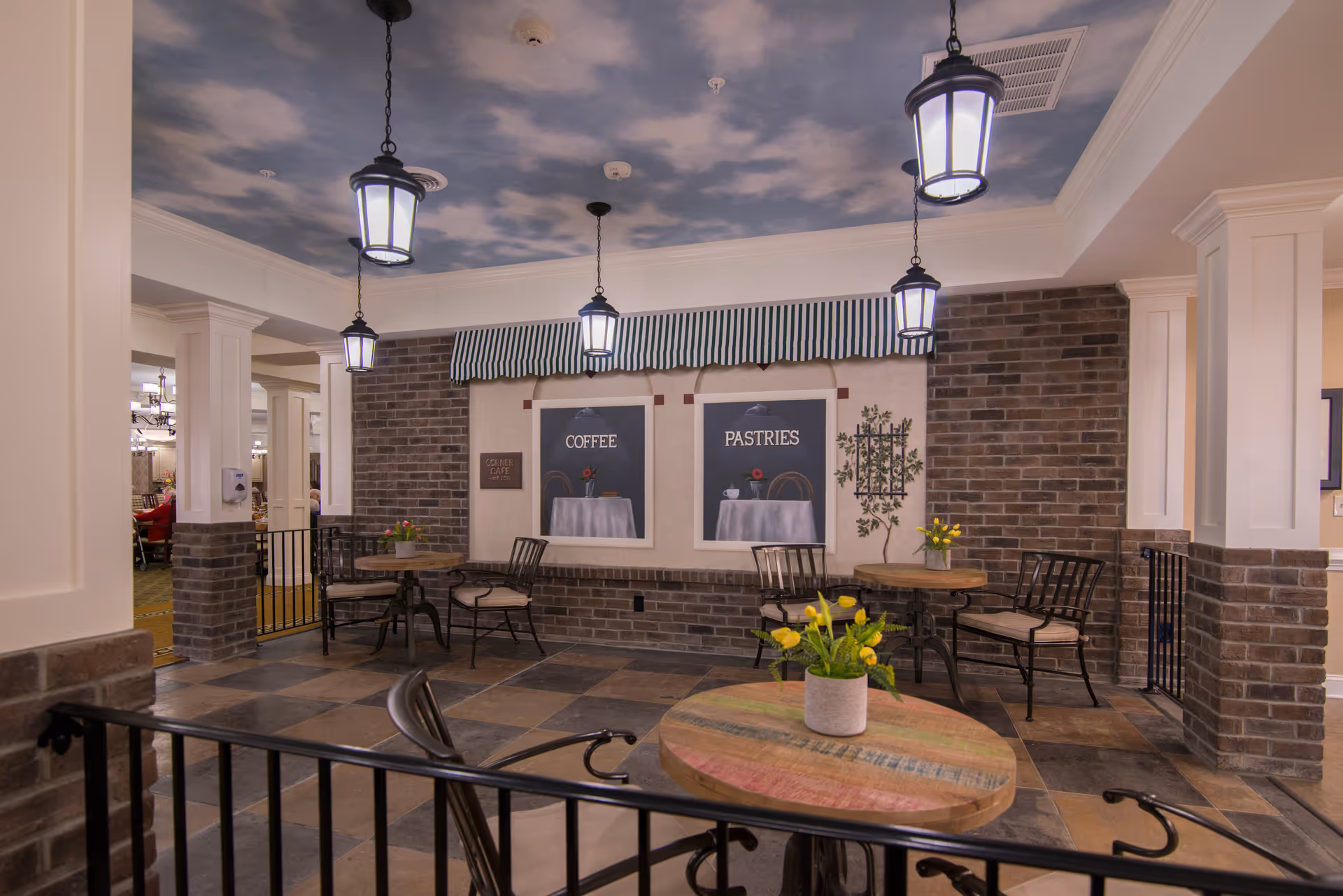 Indoor seating area with round wooden tables and metal chairs, decorated with small flower pots. The walls have a brick design with two framed signs labeled 'COFFEE' and 'PASTRIES' under a striped awning. The ceiling is painted with a sky and clouds motif and has hanging lantern-style lights.