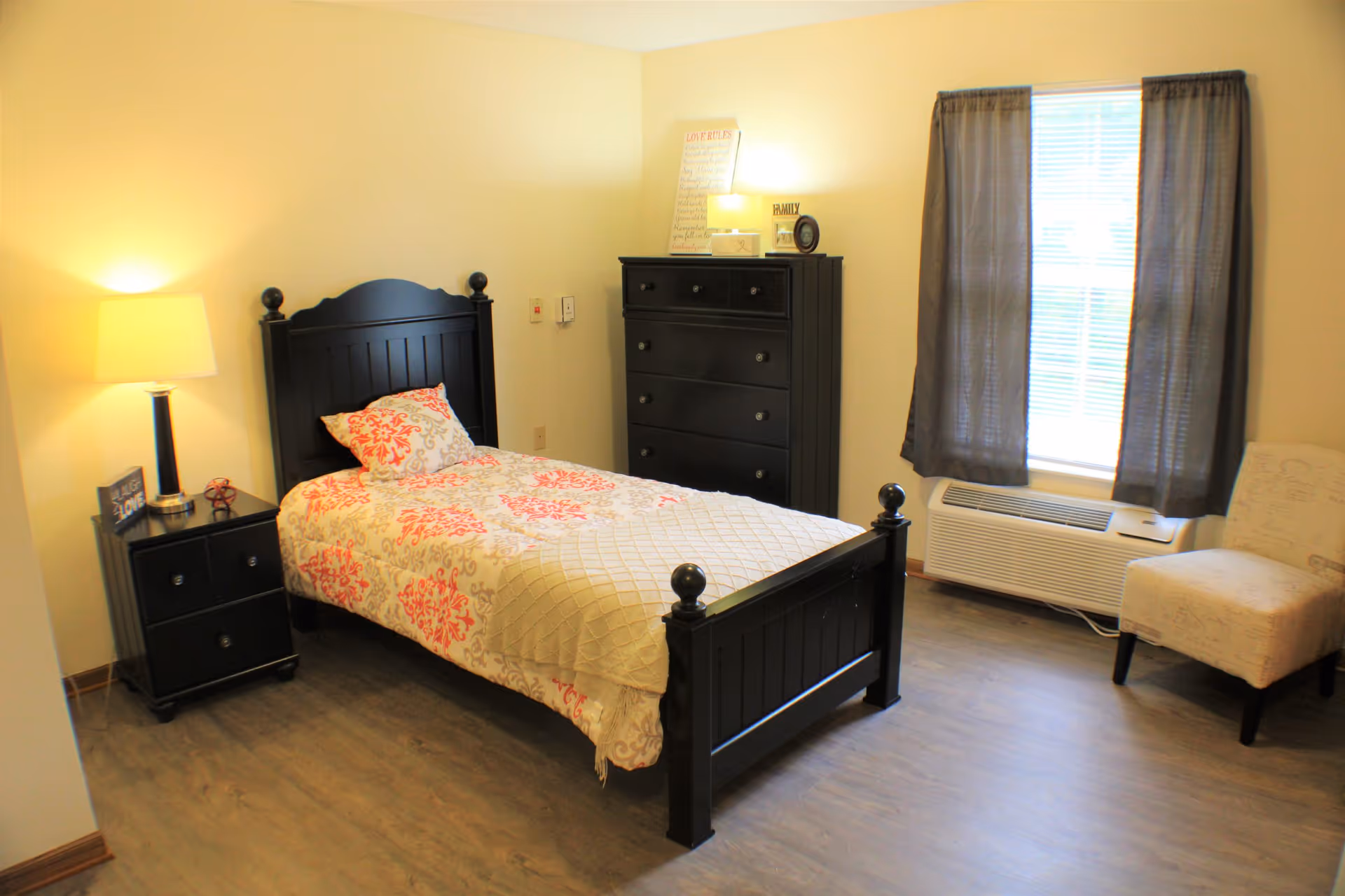A neatly arranged bedroom with a single black wooden bed featuring a patterned bedspread and pillow. Next to the bed is a black nightstand with a lamp and decorative items. Across from the bed is a black dresser with framed photos and a lamp on top. A window with dark curtains lets in natural light, and a beige upholstered chair is positioned near the window. The room has light-colored walls and wood flooring.