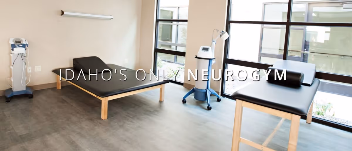 A bright therapy room with two black treatment tables, medical equipment, large windows, and overlaid text reading "IDAHO'S ONLY NEUROGYM."