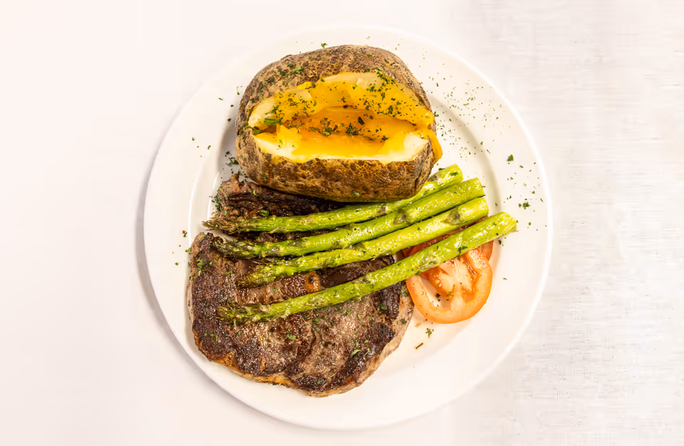 A white plate with a baked potato topped with melted cheese and herbs, grilled asparagus spears, a slice of tomato, and a grilled steak, all garnished with herbs.