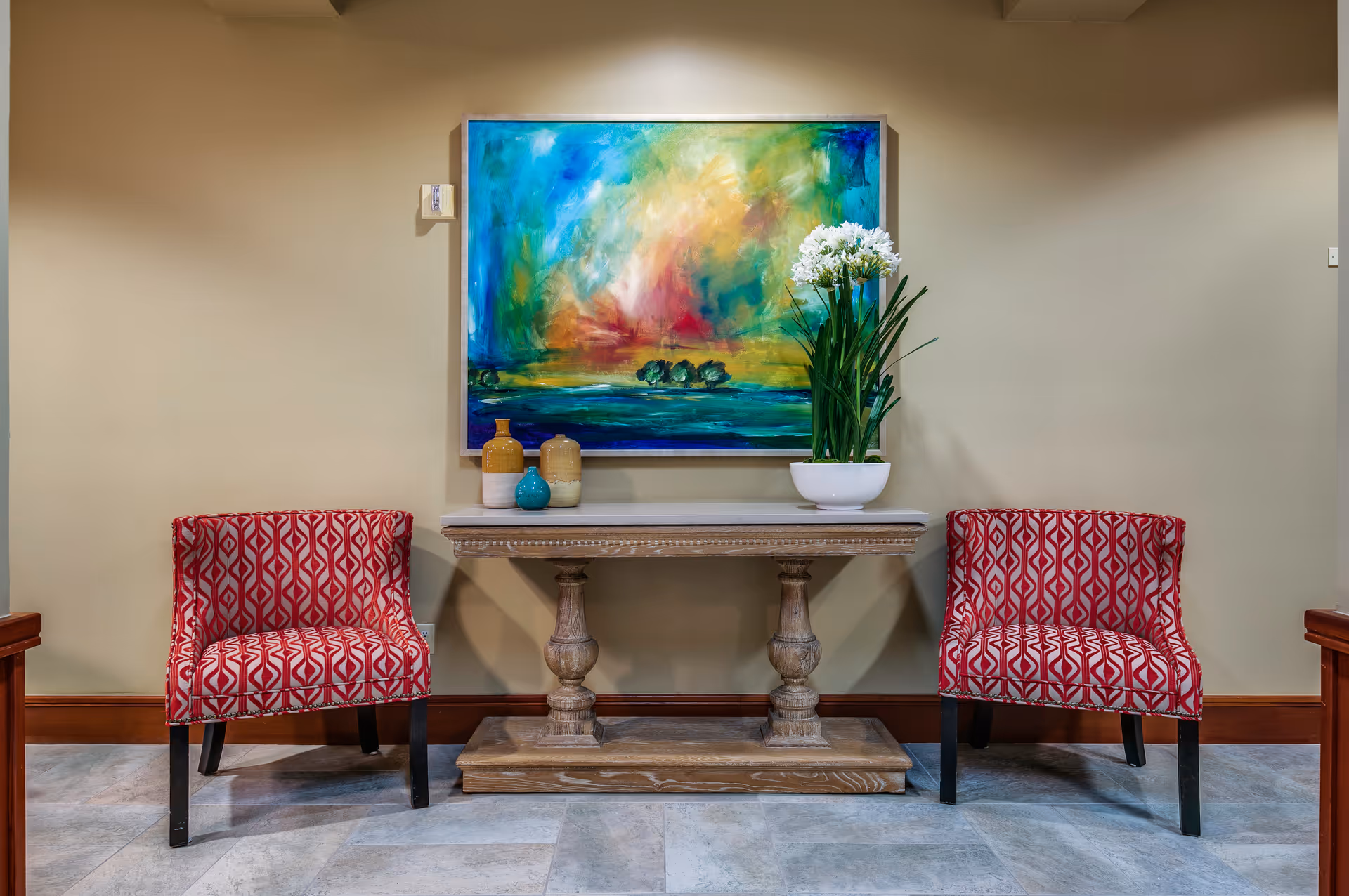 Two red patterned armchairs flank a wooden console table with decorative vases and a white bowl with white flowers. Above the table hangs a colorful abstract painting with blue, green, yellow, and red hues. The setting is indoors with beige walls and tiled flooring.