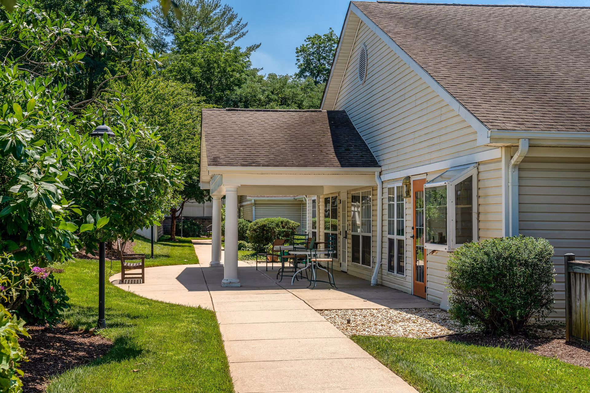 Outdoor view of a senior living facility showing a paved walkway alongside a beige building with a covered patio area. The patio has a table and chairs, surrounded by green grass, bushes, and trees under a clear blue sky.