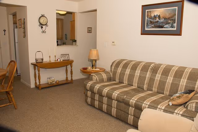 A cozy living room with a plaid patterned sofa, a small wooden side table with a lamp, a wooden console table with decorative items, and a framed painting on the wall. The room has beige carpet and light-colored walls, with a partial view into a kitchen area.