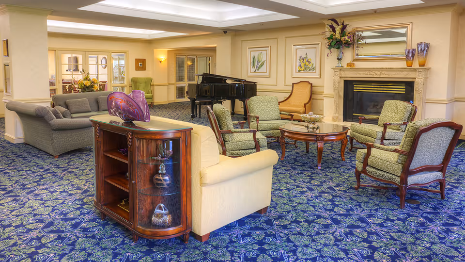 A spacious senior living community lounge with patterned blue carpet, multiple upholstered chairs and sofas arranged around a wooden coffee table, a grand piano in the background, floral artwork on the walls, and a decorative fireplace with a mirror and vases on the mantel.