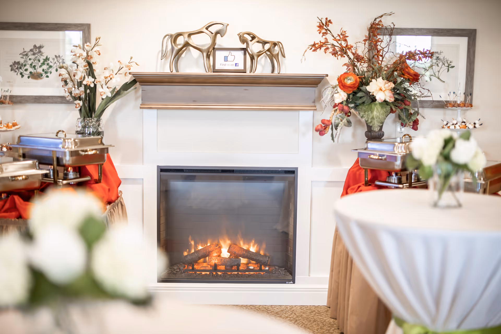 A cozy indoor setting featuring a lit fireplace with a wooden mantel decorated with two horse sculptures and a small sign. On either side of the fireplace are buffet tables draped with red and beige cloths, holding chafing dishes and floral arrangements. In the foreground, there are round tables covered with white tablecloths and small flower vases.