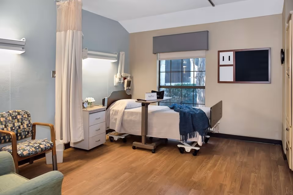 A nursing facility room with a hospital bed covered in white bedding and a blue throw blanket. The bed has an overbed table with a sign on it. There is a small white nightstand with drawers next to the bed, a chair with patterned upholstery, and a privacy curtain hanging from the ceiling. A large window with a gray valance lets in natural light. The walls are painted in soft beige and blue tones, and the floor is wood.