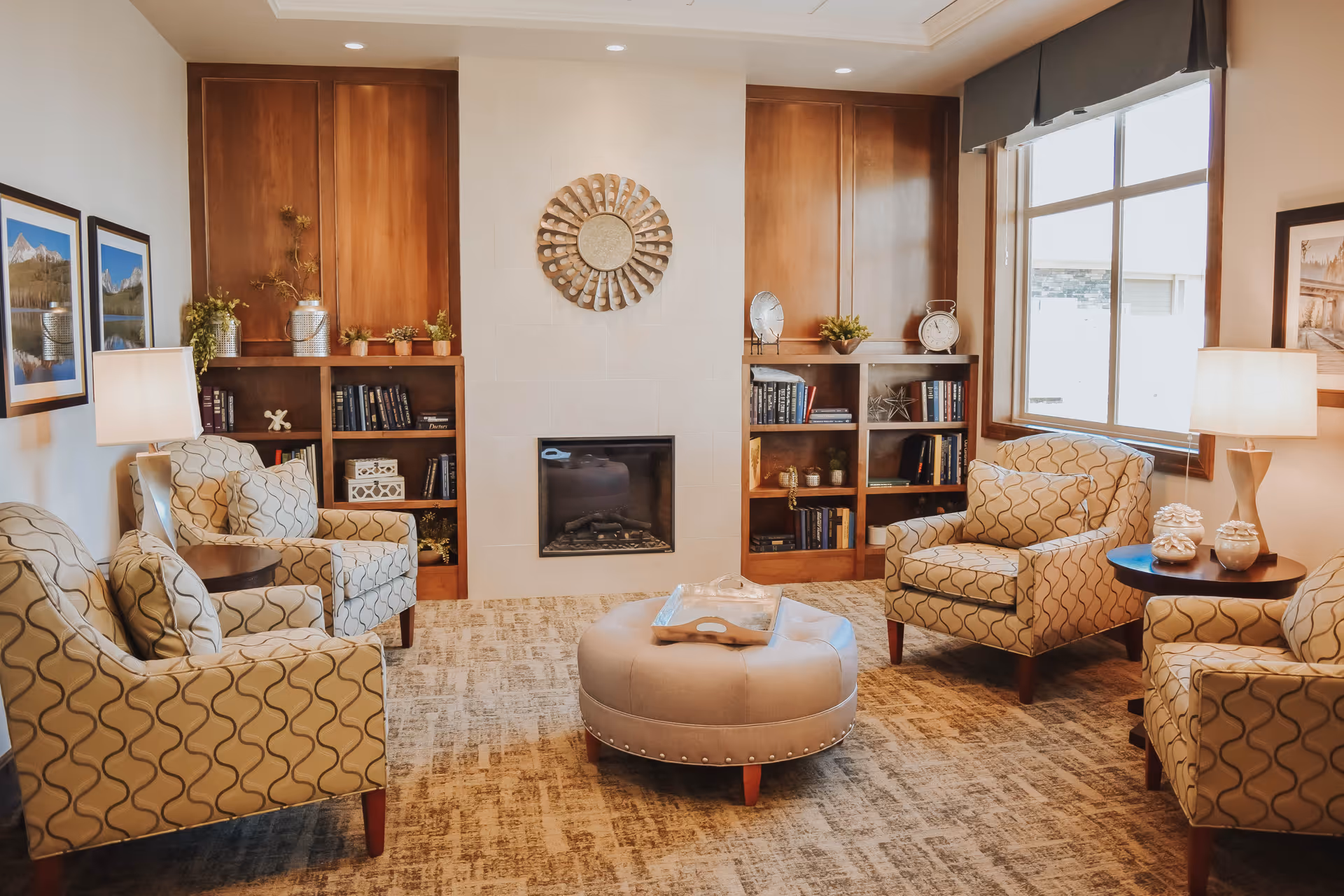 A cozy living room area with four patterned armchairs arranged around a round ottoman. The room features a fireplace centered on a wall with built-in wooden shelves on either side filled with books and decorative items. Two table lamps on side tables provide warm lighting, and large windows allow natural light to enter. The walls are decorated with framed landscape photographs.