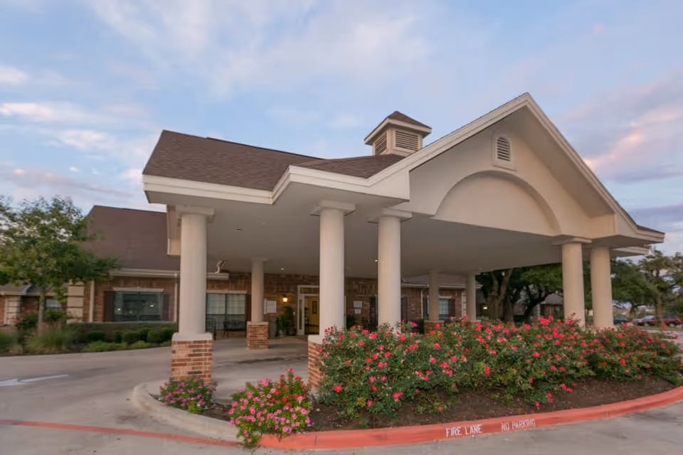 Covered entrance of a brick senior living facility with white columns, a porte-cochère, and flowering shrubs in front.