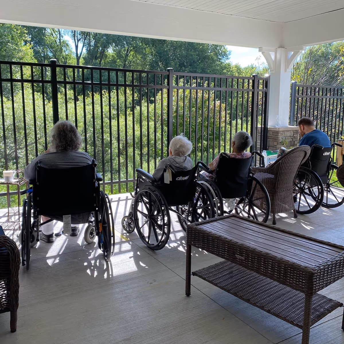 Four elderly individuals in wheelchairs sitting on a covered patio facing a black metal fence with lush green trees and bushes beyond it. The patio has wicker furniture including a table and chairs, and sunlight casts shadows on the concrete floor.