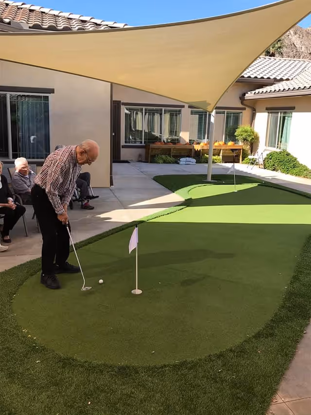 An elderly man is playing putting golf on a small artificial green in an outdoor courtyard area of a senior living facility. Several other elderly people are seated nearby watching. The courtyard is surrounded by single-story buildings with windows and has a large beige shade sail overhead providing shade.