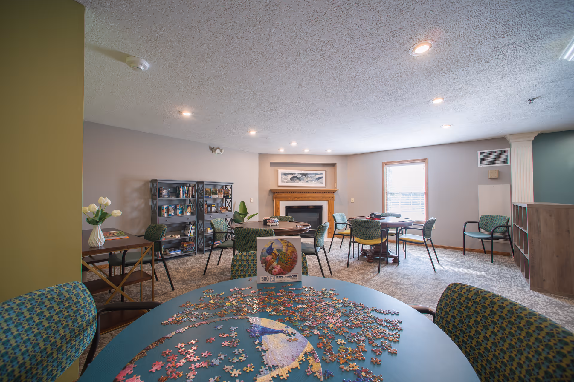 A cozy common room with multiple round tables and chairs arranged for group activities. One table in the foreground has a partially completed jigsaw puzzle with a picture of peacocks. The room features a fireplace with a framed picture above it, a window letting in natural light, bookshelves filled with books and games, and soft overhead lighting.