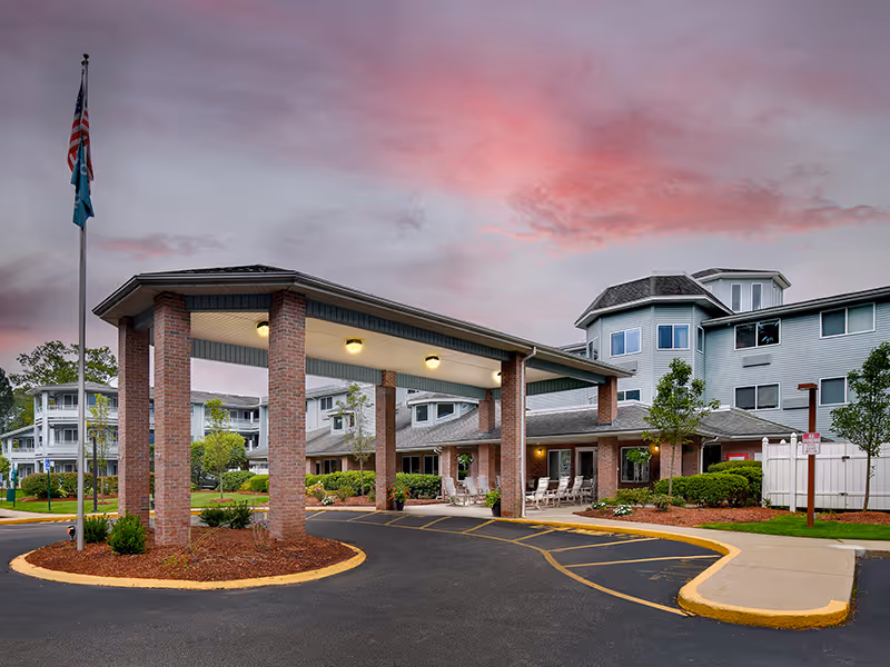 Senior living facility exterior with porte-cochere entrance, flagpole, and landscaped curbside driveway.