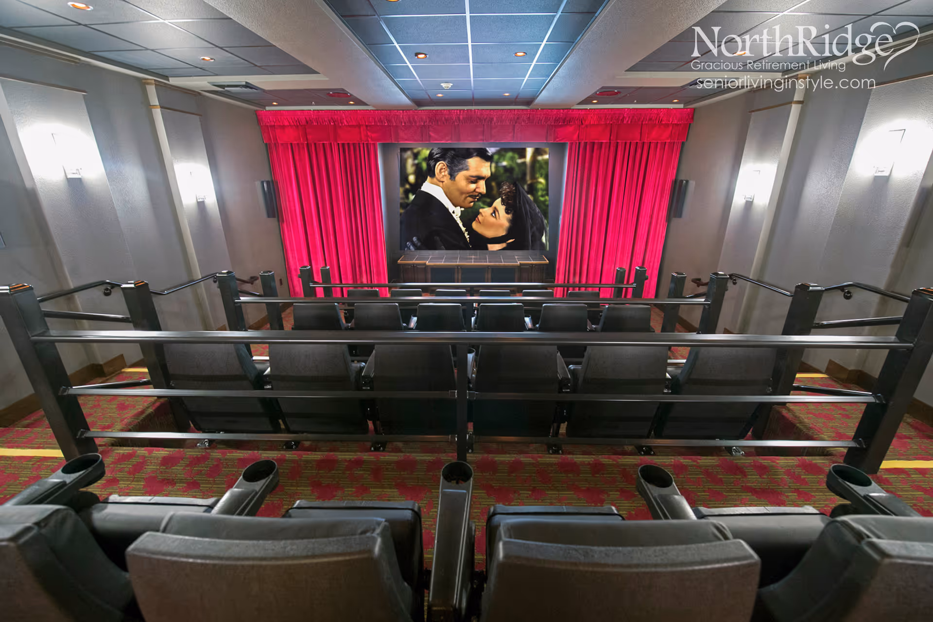 Interior view of a small movie theater room with rows of black cushioned seats facing a large screen showing a classic movie scene. The screen is framed by bright red curtains, and the room has a patterned carpet with red and yellow designs. Soft wall lights illuminate the space.