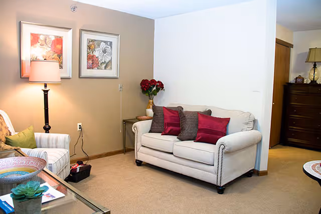 A cozy living room area with a beige loveseat adorned with red and brown throw pillows, a side table with a vase of red flowers, a floor lamp, two framed floral artworks on the wall, and a striped armchair with a green pillow. There is also a glass coffee table with decorative items and a wooden dresser in the background.