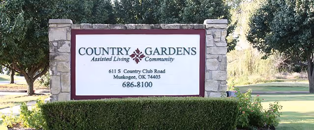 Stone entrance sign reading "Country Gardens Assisted Living Community" with address and phone number amid trees and landscaping.