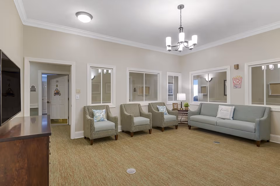 A well-lit living room with beige walls and carpeted floor featuring a light blue sofa and three matching armchairs with patterned cushions. A wooden TV stand with a flat-screen TV is on the left, and a small side table with a lamp and decorative items is next to the sofa. Several windows with white frames are on the walls, and a modern chandelier hangs from the ceiling.