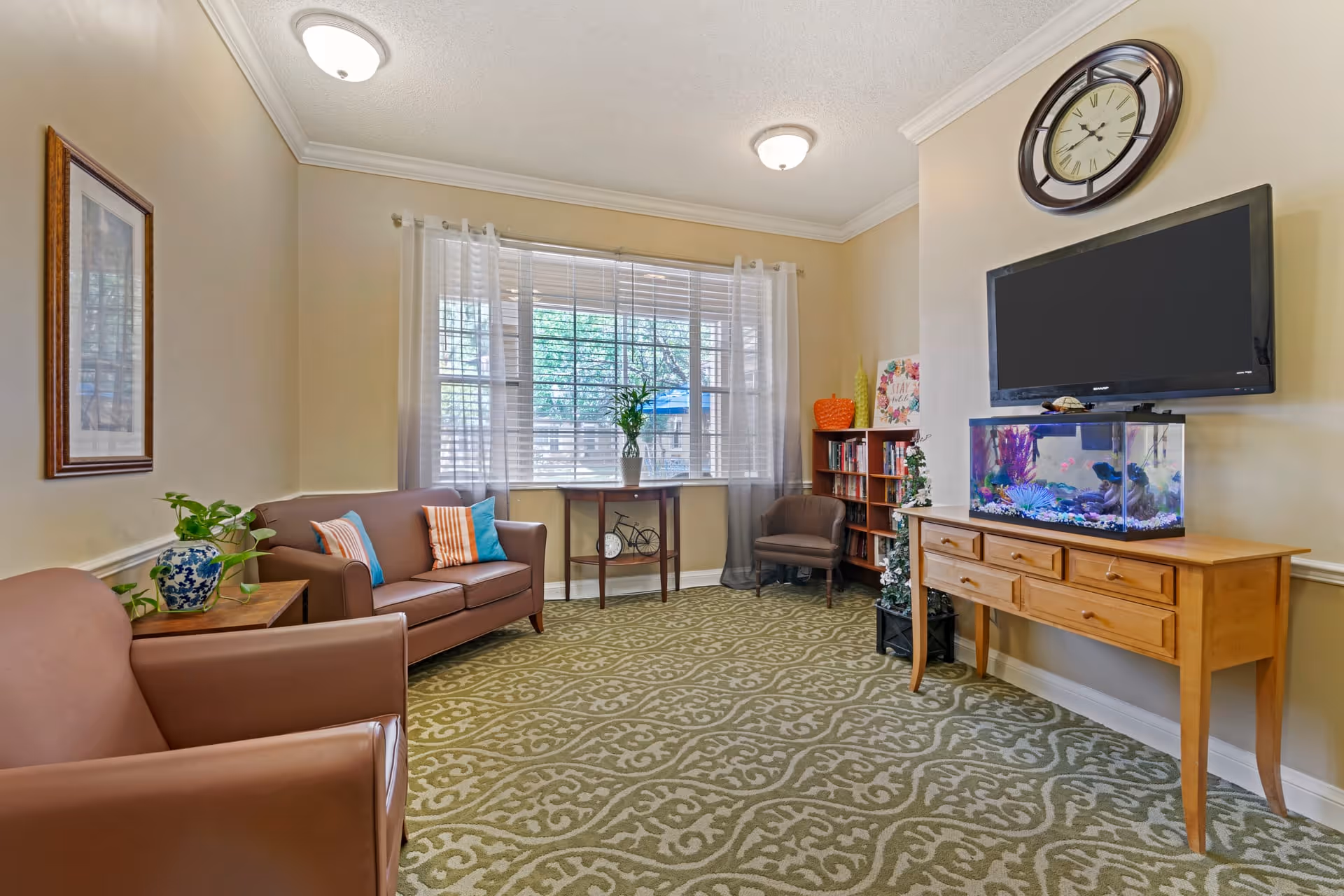 A cozy living room with beige walls and patterned green carpet. The room features two brown leather sofas with colorful pillows, a small wooden table with a plant, a window with sheer white curtains, a wooden console table with a fish tank and a flat-screen TV mounted above it, a bookshelf filled with books, and a large wall clock.