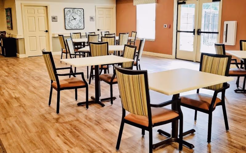 A bright dining area with several square tables and chairs arranged neatly on a wooden floor. The walls are painted in warm tones with large windows and glass doors allowing natural light to fill the room. A large clock is mounted on the wall between two doors in the background.
