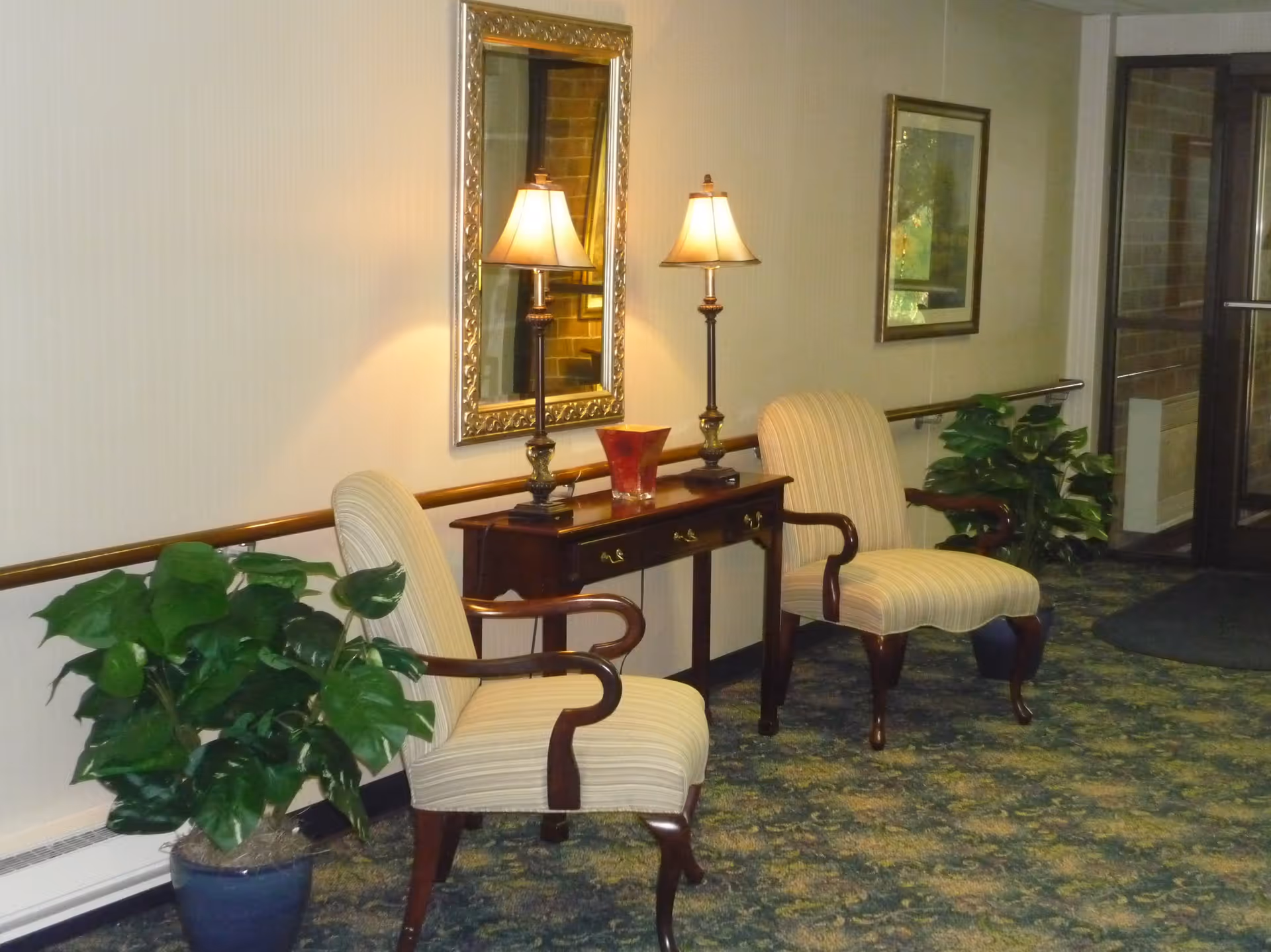 A cozy seating area in a hallway with two striped upholstered armchairs, a wooden console table with two lamps and a decorative vase, a large ornate mirror on the wall, framed artwork, and two potted plants on either side. The floor is carpeted with a patterned design and there is a glass door at the end of the hallway.