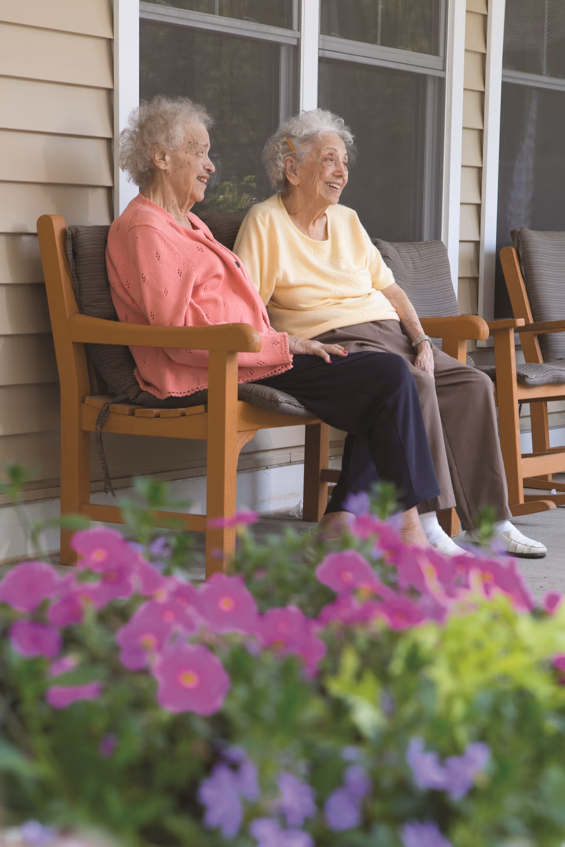 Two elderly women sitting and smiling on wooden chairs on a porch outside a building, with pink and purple flowers in the foreground.