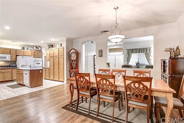 A spacious and well-lit interior featuring a dining area with a wooden table and eight matching chairs on a rug, adjacent to a kitchen with wooden cabinets, a white refrigerator, microwave, and an island. The room has hardwood floors, a chandelier above the dining table, and a grandfather clock near an arched doorway.