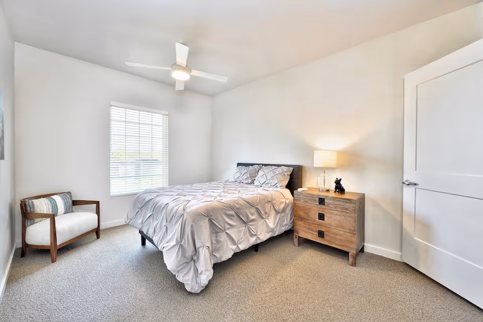Bright, minimalist bedroom with a bed, wooden nightstand and lamp, an armchair, ceiling fan, and a window with blinds.