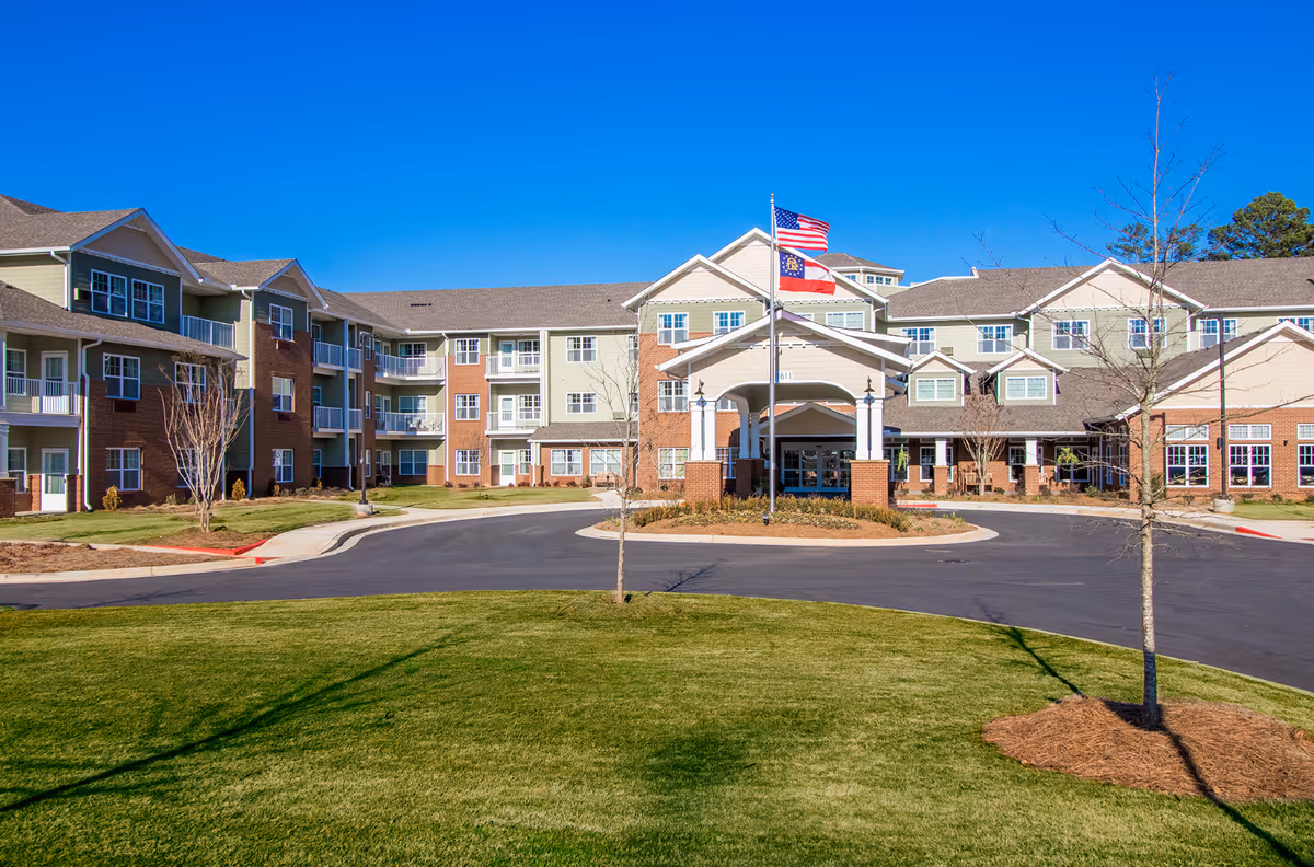 Front exterior view of Linwood Estates Gracious Retirement Living, a large multi-story building with a covered entrance, two flagpoles displaying the American and Georgia state flags, surrounded by a well-maintained lawn and young trees under a clear blue sky.