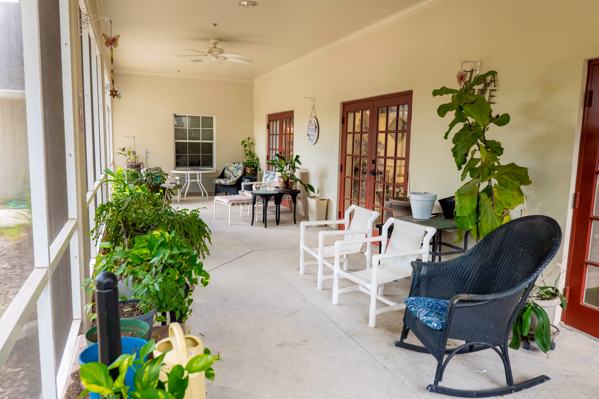 A screened covered patio with potted plants, multiple chairs and tables, and red French doors leading inside.