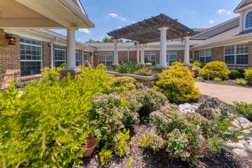 A sunny outdoor courtyard garden at Addington Place of Prairie Village featuring lush green shrubs, flowering plants, a stone-lined dry creek bed, and a pergola supported by white columns. The courtyard is surrounded by a brick building with large windows and a paved walkway.