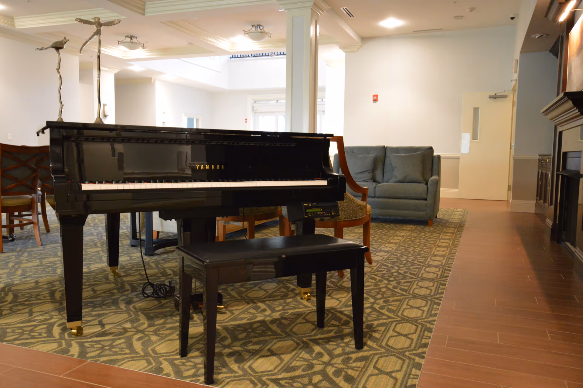 Interior of a senior living facility lounge area featuring a black Yamaha grand piano with a matching bench on a patterned carpet. Behind the piano, there are wooden chairs and a blue upholstered loveseat. The room has wooden flooring, white walls, and a fireplace on the right side.
