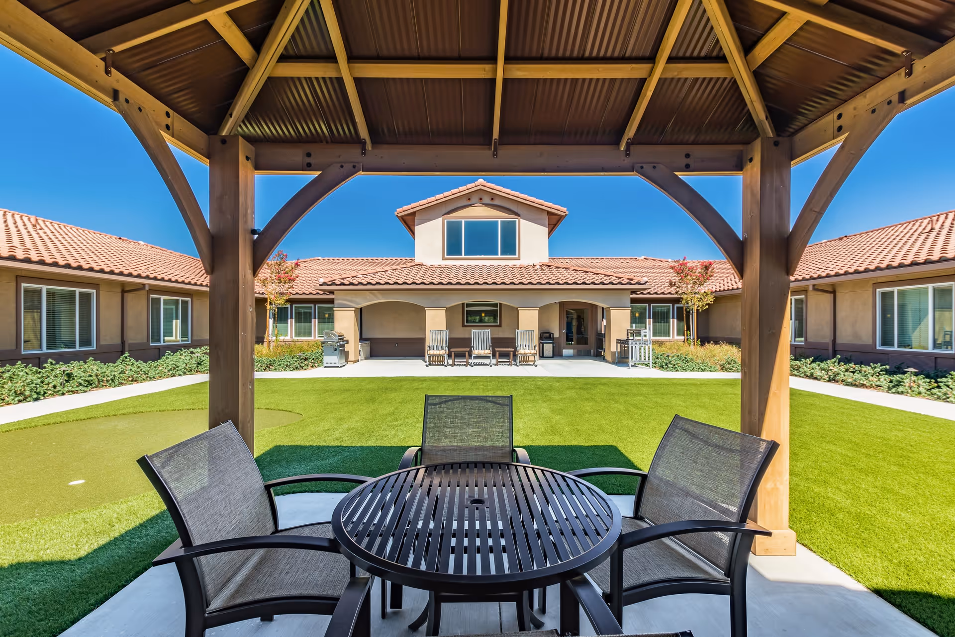 Outdoor seating area under a wooden gazebo with a round table and four chairs, overlooking a well-maintained lawn and a building with a tiled roof and multiple windows under a clear blue sky at Vineyard Place.