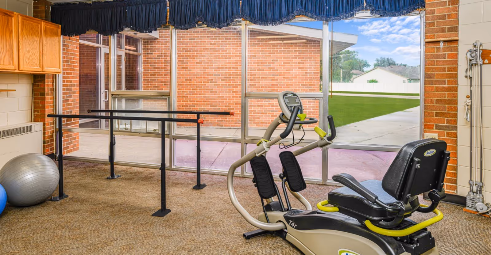 A small exercise room with a recumbent stationary bike, parallel bars for walking support, and exercise balls. The room has large windows showing an outdoor view with a brick wall and green lawn outside.