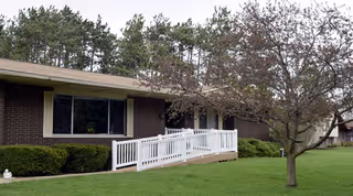 Exterior view of a single-story brick building with a white wheelchair ramp leading to the entrance. There is a tree with sparse leaves on the right side and a well-maintained green lawn in front.