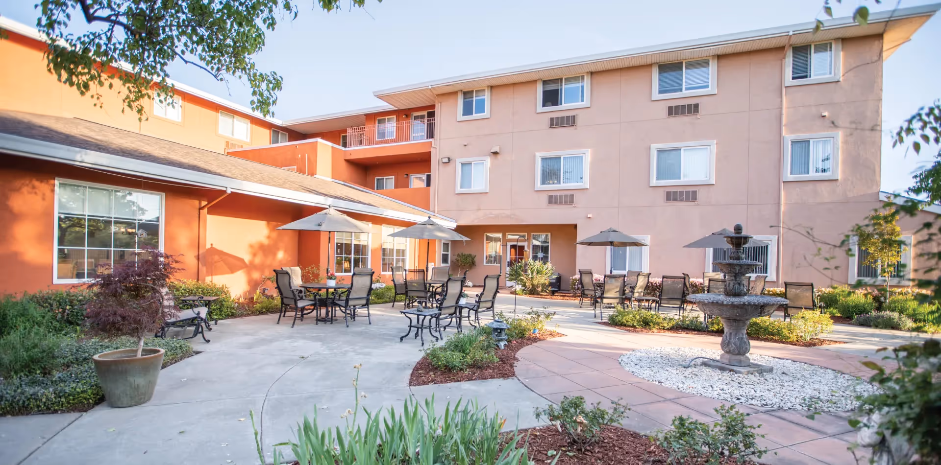 Courtyard patio with tables, umbrellas, chairs and a fountain in front of a three-story residential building.