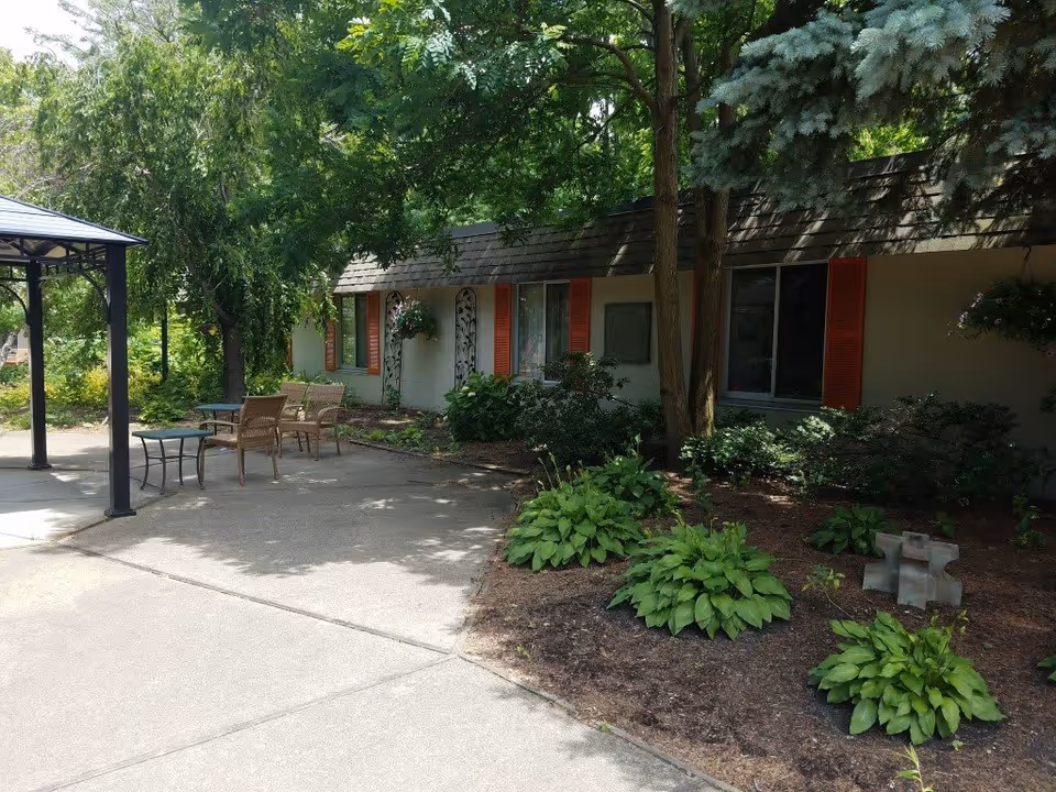 Outdoor patio area with a concrete walkway, several chairs and a small table under a metal gazebo. The area is surrounded by greenery including bushes, trees, and plants. A building with beige walls and red window shutters is visible in the background.