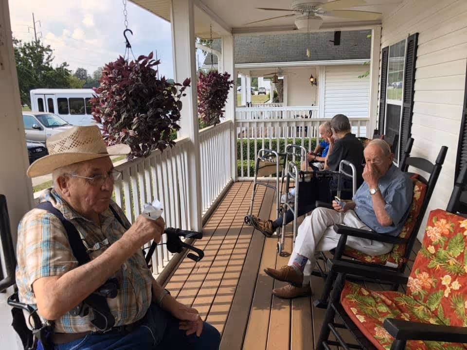Four elderly men sitting on a covered porch with wooden flooring and white railing. They are seated on rocking chairs with floral cushions, some using walkers. One man in the foreground is wearing a straw hat and plaid shirt, holding a small object. Hanging plants are visible along the porch railing, and a white building and parked vehicles can be seen in the background.