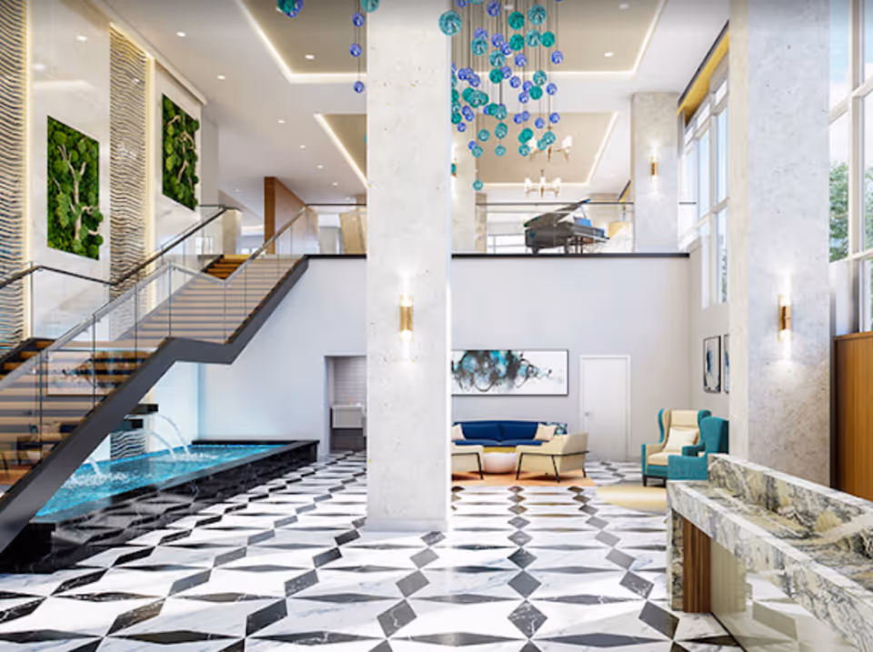 A spacious and modern senior living facility lobby with a black and white geometric tiled floor, a water feature with cascading water under a staircase, comfortable seating areas with blue and beige chairs, large windows letting in natural light, green wall art, and a grand piano on the upper level.