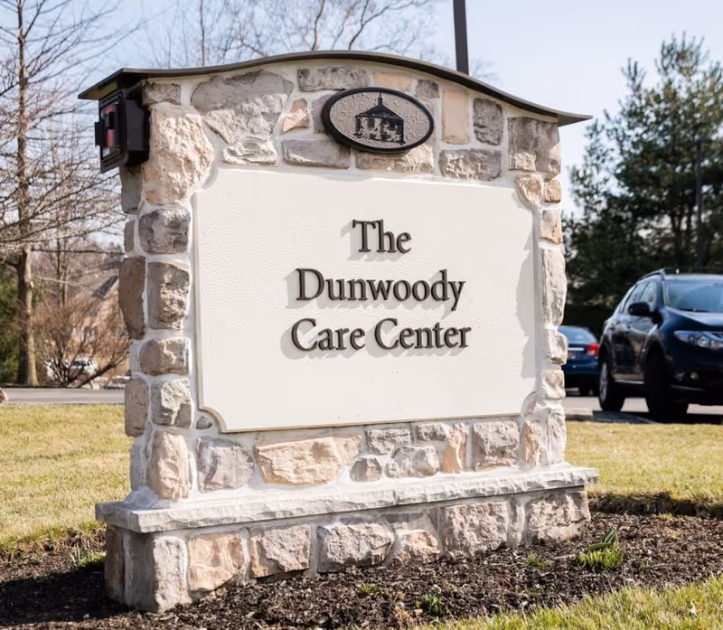 Stone monument sign with a white plaque that reads 'The Dunwoody Care Center' situated on a grassy area with trees and parked cars in the background.