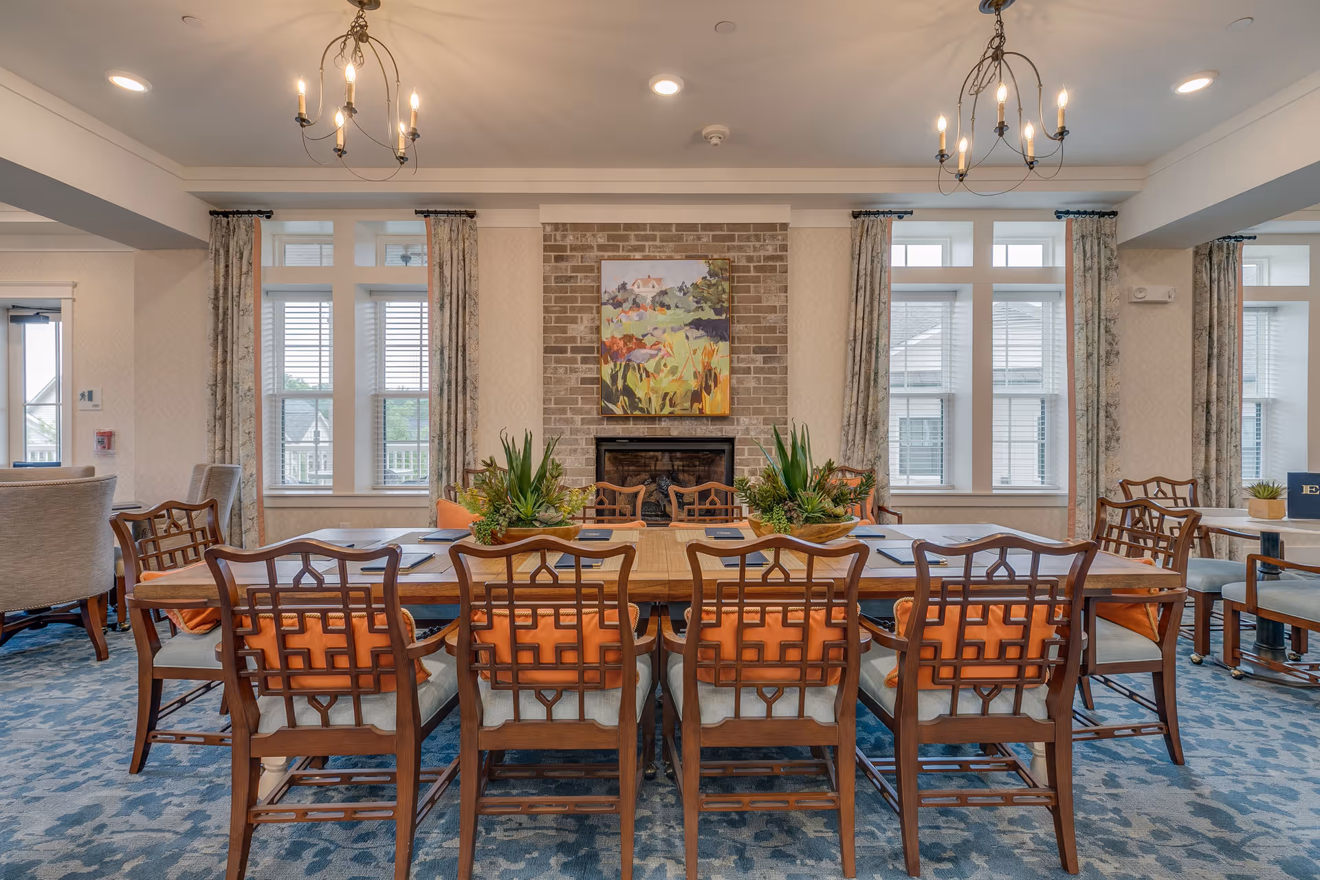 Bright communal dining room with a long wooden table, multiple chairs, a fireplace, and large windows.