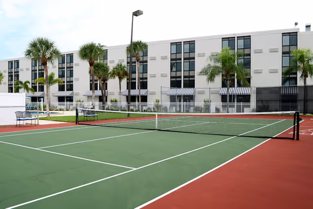 Tennis court with a net in front of a multi-story senior living building flanked by palm trees and striped awnings.