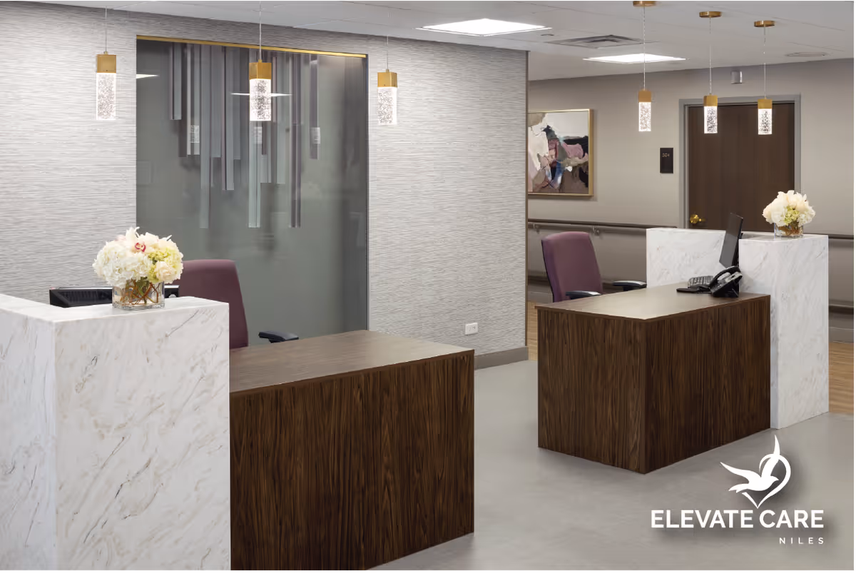 Reception area with two wooden desks featuring white marble accents, purple office chairs, pendant lights hanging from the ceiling, and floral arrangements on the desks. A hallway with a door and a framed abstract painting is visible in the background.