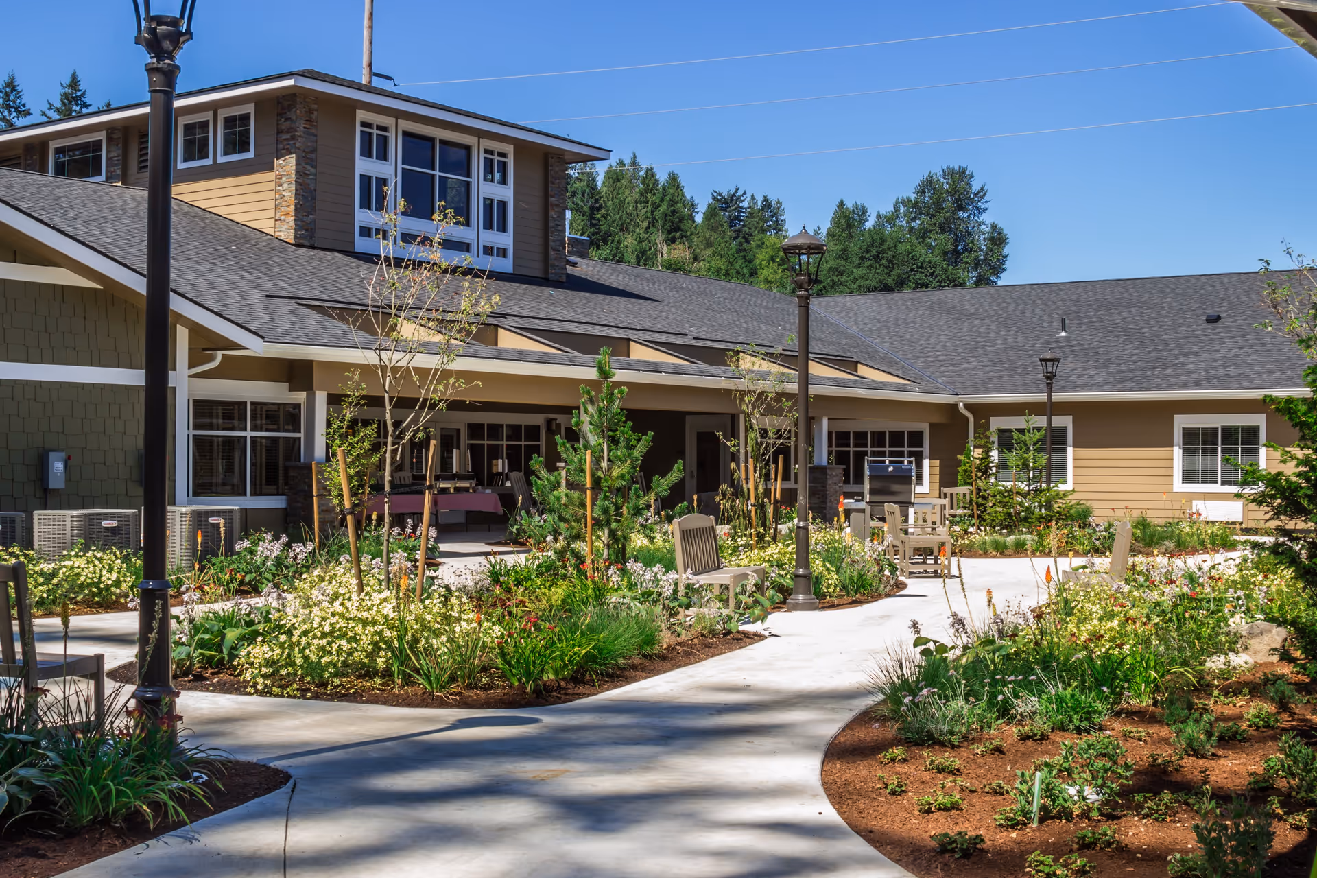 Outdoor courtyard area of a senior living facility with a paved walkway, garden beds filled with flowers and shrubs, benches, and lampposts. The building surrounding the courtyard has beige siding, multiple windows, and a gray roof under a clear blue sky.
