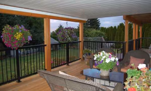 Outdoor balcony with wicker seating, potted flowers, and hanging baskets overlooking a garden.