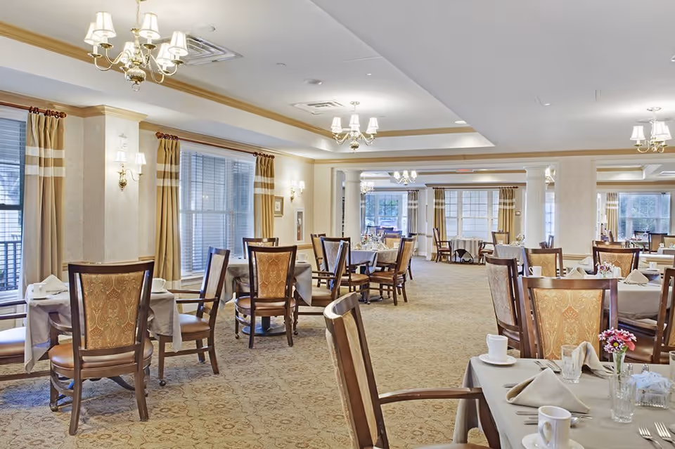 A spacious and well-lit dining room with multiple tables covered in beige tablecloths, each set with cups, glasses, napkins, and small flower arrangements. The room features large windows with beige curtains, elegant chandeliers, and wall sconces providing warm lighting. The carpet has a subtle patterned design, and the chairs have wooden frames with upholstered backs and seats.
