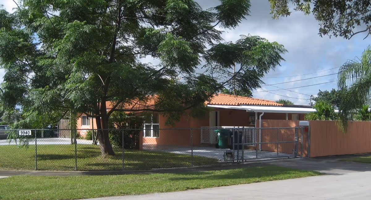 Exterior view of a single-story building with a red tile roof, surrounded by a chain-link fence and trees. The building has a driveway with trash bins and a wooden privacy fence on one side. The address number 9940 is visible on the fence.