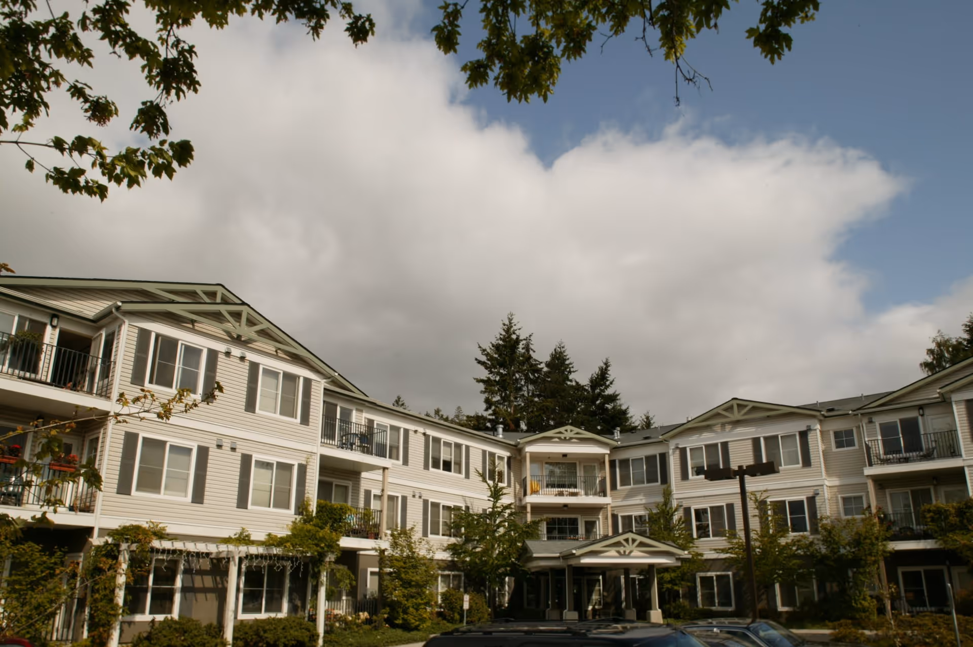 Exterior view of a three-story senior living facility building with balconies, surrounded by trees and greenery under a partly cloudy sky.